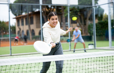 Wall Mural - Emotional young European girl padel player hitting ball with racket on hard court in autumn