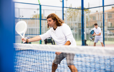 Wall Mural - Focused middle-aged Latin man padel player hitting ball with racket on hard court in summer