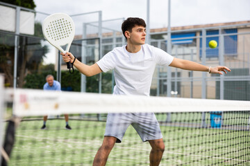 Wall Mural - Portrait of emotional determined young guy playing padel tennis on open court in summer, swinging racket to return ball over net. Sportsman ready to hit volley