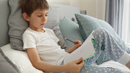 Portrai of little boyworking on his homework in bed, writing in a notebook with concentration. Importance of education and child development in a remote learning environment.