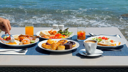 Wall Mural - Delicious buffet breakfast served with a variety of eggs, sausage, coffee, fresh orange juice, croissants, and salad with sea view. Man enjoys his meal, dining outside, enjoying idyllic ocean view.