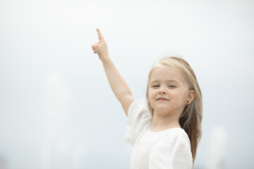 Wall Mural - Look there. Smiling little girl on the sky background pointing up with her finger. Focus at her face. Copyspace for your text or logo, banner