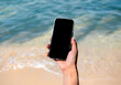 © tete_escape - Mobile beach phone with empty screen. Mockup image of black blank desktop screen in hand holding mobile smart phone on the blue sea background in summer. Travel booking, searching, business marketing.