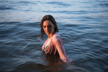 Beautiful caucasian woman in pink dress in water on the sunset