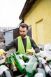 © LIGHTFIELD STUDIOS - Bearded male worker in protective vest and gloves standing near blurred glass trash in outdoor waste disposal station, garbage sorting and recycling concept