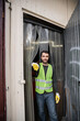 © LIGHTFIELD STUDIOS - Cheerful male worker in fluorescent vest and protective gloves looking at camera while standing near door of waste disposal station, garbage sorting and recycling concept