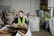 © LIGHTFIELD STUDIOS - Young female worker in protective vest and gloves putting waste paper in sack while working near blurred indian colleague in waste disposal station, garbage sorting and recycling concept