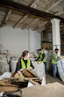 © LIGHTFIELD STUDIOS - Young female worker in protective gloves and vest looking at multiethnic colleagues while putting cardboard in sack in waste disposal station, garbage sorting and recycling concept