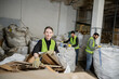 © LIGHTFIELD STUDIOS - worker in gloves and high visibility vest looking at camera while putting cardboard in sack and working near multiethnic colleagues in waste disposal station, garbage sorting and recycling concept