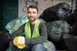 © LIGHTFIELD STUDIOS - Smiling and bearded worker in safety vest and gloves looking at camera while sitting on plastic bags with trash in blurred garbage sorting center, recycling concept