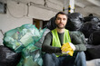© LIGHTFIELD STUDIOS - Bearded sorter in protective vest and gloves looking at camera while sitting near plastic bags with trash while working in blurred garbage sorting center, recycling concept