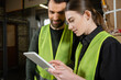 © LIGHTFIELD STUDIOS - Young worker in safety reflective vest using digital tablet while standing near smiling colleague and blurred waste paper in garbage sorting center, waste sorting and recycling concept