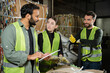 © LIGHTFIELD STUDIOS - Smiling worker in reflective vest and gloves talking to multiethnic colleagues with digital tablet and standing near waste paper on hand pallet truck in garbage sorting center, waste recycling