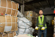© LIGHTFIELD STUDIOS - Smiling and bearded worker in reflective vest and gloves using hand pallet truck while moving waste paper in blurred garbage sorting center, waste sorting and recycling concept