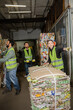 © LIGHTFIELD STUDIOS - Smiling female worker in reflective vest and gloves pointing away while standing near waste paper and colleagues with hand pallet truck in garbage sorting center, waste sorting and recycling concept