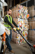 © LIGHTFIELD STUDIOS - Bearded male worker in protective vest and gloves using hand pallet truck while standing near waste paper in garbage sorting center, waste sorting and recycling concept