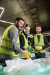© LIGHTFIELD STUDIOS - Cheerful young worker in high visibility vest and gloves standing near multiethnic colleagues near sacks in blurred waste disposal station, garbage sorting process