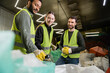 © LIGHTFIELD STUDIOS - Smiling workers in high visibility vests standing near indian colleague in protective glove holding trash near sacks while working together in blurred waste disposal station, garbage recycling