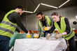 © LIGHTFIELD STUDIOS - Young worker in reflective vest and gloves holding plastic containers near sack while sorting trash with multiethnic colleagues in waste disposal station, garbage sorting and recycling concept