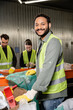 © LIGHTFIELD STUDIOS - Cheerful indian sorter in protective gloves and safety vest looking at camera while standing near plastic bag, blurred conveyor and colleagues working in waste disposal station, recycling concept