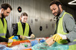 © LIGHTFIELD STUDIOS - Bearded indian worker in safety vest and protective gloves putting paper cup in plastic bag while separating trash near conveyor and colleagues in waste disposal station, recycling concept