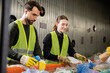 © LIGHTFIELD STUDIOS - Smiling young worker in protective gloves and safety vest separating trash on conveyor while working with colleague together in waste disposal station, recycling concept