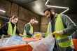 © LIGHTFIELD STUDIOS - Indian worker in protective vest and gloves putting plastic trash in bag while working near blurred colleagues and conveyor in waste disposal station, garbage sorting and recycling concept