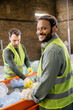 © LIGHTFIELD STUDIOS - Positive indian worker in protective clothes looking at camera while standing near trash on conveyor and blurred colleague in garbage sorting center, garbage sorting and recycling concept