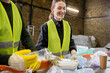 © LIGHTFIELD STUDIOS - Positive young worker in gloves and protective vest holding plastic trash near blurred colleague and conveyor in waste disposal station, garbage sorting and recycling concept