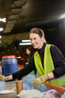 © LIGHTFIELD STUDIOS - Cheerful young worker in protective vest and gloves sorting garbage on conveyor and looking away while working in waste disposal station, garbage sorting and recycling concept