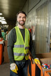 © LIGHTFIELD STUDIOS - cheerful sorter in high visibility vest and protective gloves looking at camera while standing near plastic caps in carton boxes in waste disposal station, garbage sorting and recycling concept
