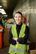 © LIGHTFIELD STUDIOS - positive and young worker in gloves and high visibility jacket crossing arms and looking at camera while standing in blurred waste disposal station at background, garbage sorting and recycling concept
