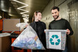 © LIGHTFIELD STUDIOS - Smiling volunteer holding trash bag near man with bin and recycle sign in blurred waste disposal station at background, garbage sorting and recycling concept