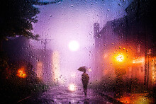 View Through A Glass Window With Raindrops On A Blurred Silhouette Of A Girl With Umbrella Walking On Autumn Rain , Night Street Scene. Focus On Raindrops