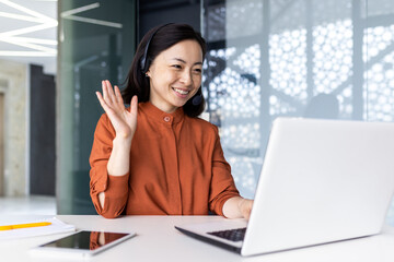 Happy Asian woman working inside office, businesswoman with video call headset talking and advising customers remotely, tech support online store customer service, greeting with hand.