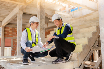 Wall Mural - Two Asian male engineers are spreading blueprints and helping each other check the work, smiling and having fun at the architectural construction site. For future industries, wear vests and helmets.