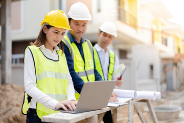 Wall Mural - Female engineer working with laptop. Two male engineers nearby working in architecture construction site. About the business industry wearing vests, helmets, tablets, blueprints and walkie-talkies.