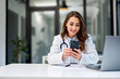 © bnenin - A female doctor sitting at the office table in front of the laptop and using a mobile phone.
