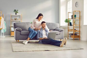 Young married couple sitting on sofa in the living room at home using tablet PC for internet and social media. Happy family resting on couch enjoying weekend watching video or talking on video call.