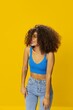© SHOTPRIME STUDIO - Woman with curly afro hair in a blue T-shirt on a yellow background dancing flying hair with sunglasses yellow, hand signs, look into the camera, smile with teeth and happiness, copy space