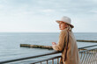 © Sergio - Stylish pensive mature woman in brown round hat and coat standing on embankment and looking away at sea horizon. Lonely elegant senior lady in nature, copy space