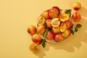 Wall Mural - Colorful fresh ripe peaches (prunus persica) with green leaves on the stem are placed on a round plate and decorated on a yellow background. Minimal scene with copy space