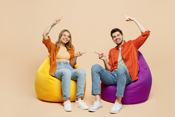 Wall Mural - Full body young couple two friends family man woman wear casual clothes together sit in bag chair point index finger between them on area isolated on pastel plain light beige color background studio.