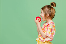 SIde View Little Child Kid Girl 6-7 Years Old Wear Casual Clothes Have Fun Hold In Hand Biting Red Apple Isolated On Plain Pastel Green Background Studio. Mother's Day Love Family Lifestyle Concept.