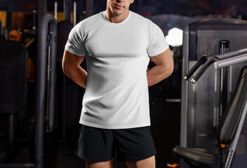 Wall Mural - White sports t-shirt mockup on a sports guy with hands behind his back, shirt on an athlete in the gym, front view.