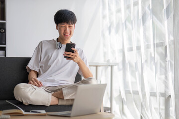 A happy young Asian man in casual clothes is looking out the window and daydreaming about his happiness while sitting on a sofa with his phone in his hands.