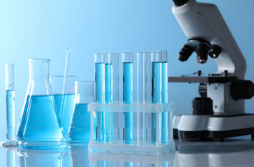 Microscope near different laboratory glassware and test tubes with light blue liquid on table