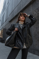 Wall Mural - Fashionable young beautiful girl in fashion urban rock outfit with black leather jacket and sweater with bag walks in the city near a black modern building