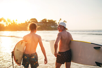 Wall Mural - Father with teenager son walking with surfboards by sandy ocean beach with palm trees on background lightened with sunset sun They have conversation and enjoy summertime Family active vacation concept