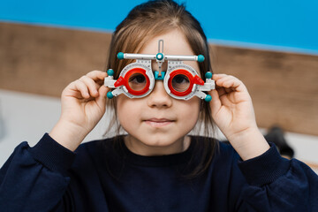 Child in trial frame glasses on blue background. Examination with pediatric ophthalmologist for selection of trial glasses frame to examine eye visual system.